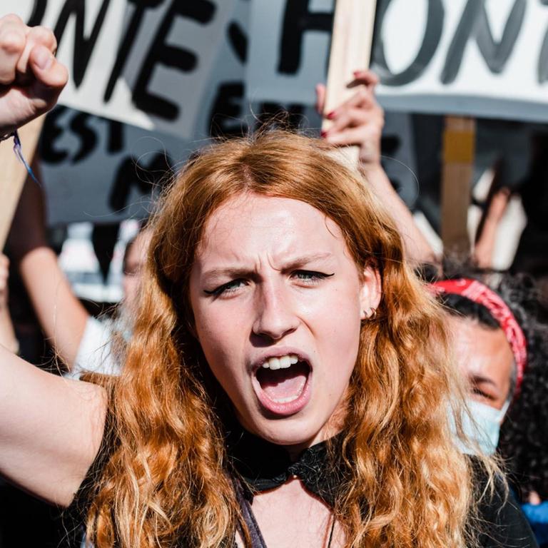 Frauendemonstration in Paris: Im Mittelpunkt des Bildes eine junge Frau mit rotblonden langen Haaren, die eine Faust in die Höhe gereckt hat. Im Hintergrund weitere in die Höhe gereckte Fäuste und Plakate.