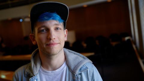 EIn junger Mann mit blauen Haaren, er trägt Jeansjacke, T-Shirt und Käppi, in einem Veranstaltungssaal. Es handelt sich um den Youtuber Rezo.