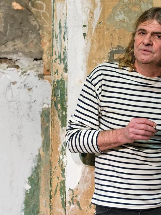 Der Musiker Wenzel mit Zigarette in der Hand vor einer heruntergekommenen Wand