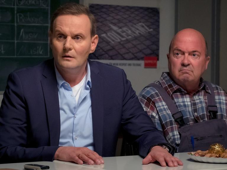 "Das Unwort": Direktor Stege (Devid Striesow) und Hausmeister Eichmann (Florian Martens) sitzen nebeneinander an einem der Schultische. Vor ihnen steht ein Teller mit Leckereien, im Hintergrund sind eine Kreidetafel sowie ein Plakat mit der Aufschrift "Toleranz" zu erkennen. Stege legt beide Hände flach auf den Tisch und schaut entsetzt. Eichmann blickt mürrisch in dieselbe Richtung wie Stege.