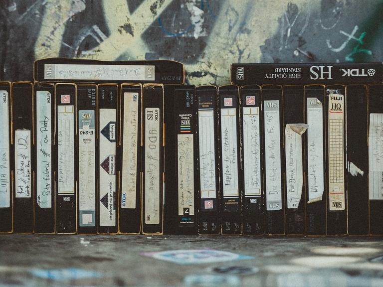 Eine Reihe VHS-Kassetten stehen vor einer mit Graffiti bemalten Wand