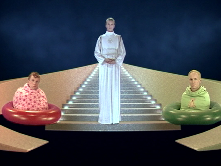Videostill vom Künstler Bjørn Melhus, eine priesterliche Figur steht in der Mitte auf einer Treppe, daneben zwei Menschen in Schwimmreifen