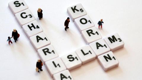 Mit Buchstabenplättchen sind die Worte "Scharia", "Islam" und "Koran" gelegt. Kleine Menschen-Figuren stehen um die Buchstaben herum