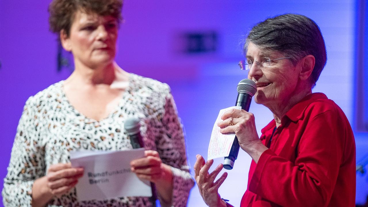 Moderatorin und Historikerin stehen mit Mikrophonen auf einer lila ausgeleuchteten Bühne nebeneinander.