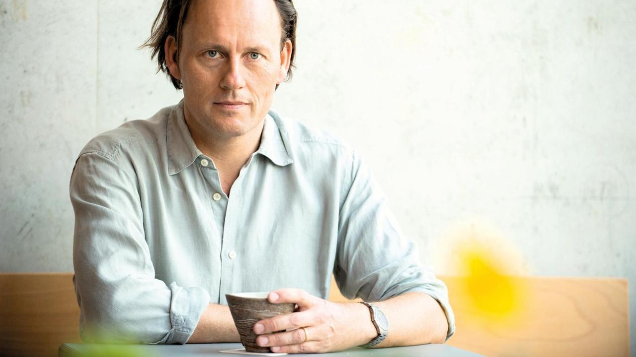 Der Autor Gregor Hens sitzt in grauen Hemd vor einer grauen Wand an einem Tisch, in der Hand eine Tasse.