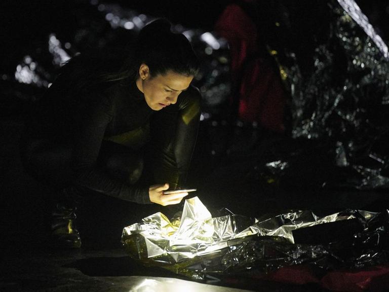 Eine Frau steht in dunkler Umgebung mit ihrem Smartphone in der Hand gebückt über einem Gebilde aus Alufolie, das hell erleuchtet ist.