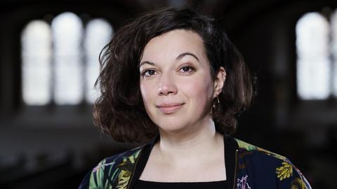 Die Pastorin Jasmin El-Manhy steht in einer Kirche und schaut in die Kamera. Im Hintergrund sind die Kirchenfenster zu sehen.