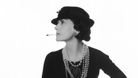 Ein Schwarzweiß-Foto zeigt die französische Modemacherin Gabrielle Coco Chanel im Profil in schwarzen Kleid mit Halsketten und Zigarette im Mund