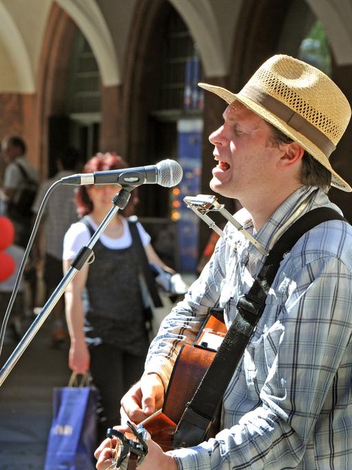 Der Musiker Jimmy Kelly singt im Stadtzentrum von Leipzig als Straßenmusikant. Er trägt einen Strohsonnenhut und spielt Gitarre.