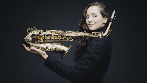 Eine Frau mit langen braunen Haaren hält ein Saxofon in der Hand. Sie balanciert es auf der linken Schulter und der ausgetreckten linken Hand. Es ist die Saxofonistin Asya Fateyeva.