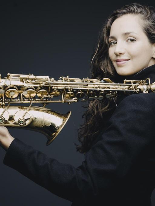 Eine Frau mit langen braunen Haaren hält ein Saxofon in der Hand. Sie balanciert es auf der linken Schulter und der ausgetreckten linken Hand. Es ist die Saxofonistin Asya Fateyeva.