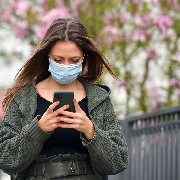 Eine junge Frau mit Schutzmaske blickt auf ihr Smartphone. (Symbolbild)