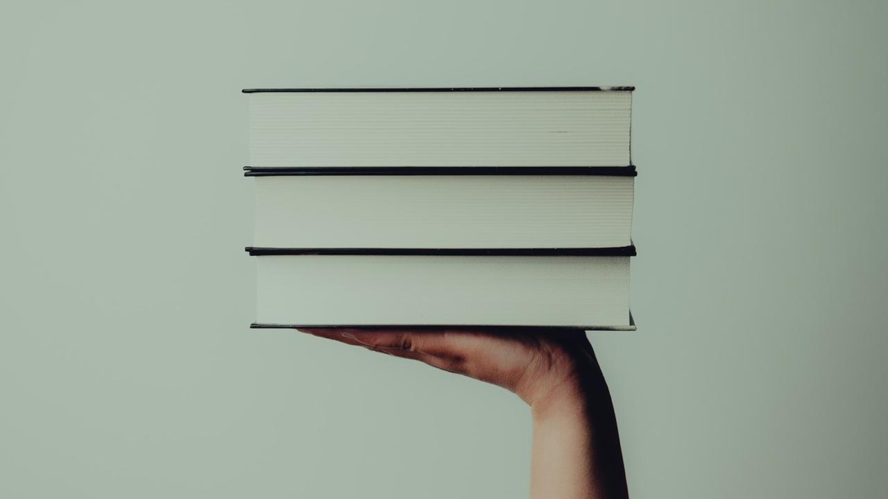 Eine Hand hält vor mintgrünem Hintergrund drei Bücher hoch.