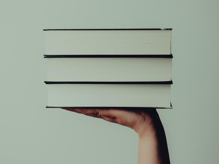 Eine Hand hält vor mintgrünem Hintergrund drei Bücher hoch.