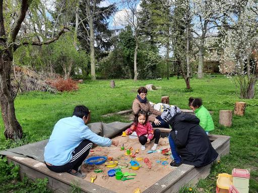 In einem großen Garten mit zahlreichen Bäumen steht ein Sandkasten, in dem zwei kleine dunkelhaarige Mädchen spielen. Auf dem Rand sitzen die Eltern und eine weitere Frau.