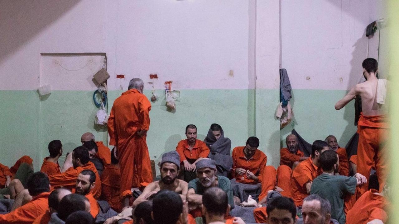 Viele Männer in roten Overalls sitzen eng zusammengepfercht in einem großen Raum mit grün-weißen Wänden.