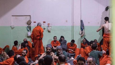 Viele Männer in roten Overalls sitzen eng zusammengepfercht in einem großen Raum mit grün-weißen Wänden.