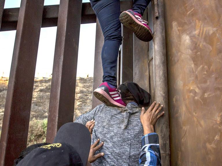 Migranten aus Mexiko klettern über den Grenzzaun in die USA. Füsse stehen auf einer Schulter und Hände werden sichernd in die Luft gereckt.