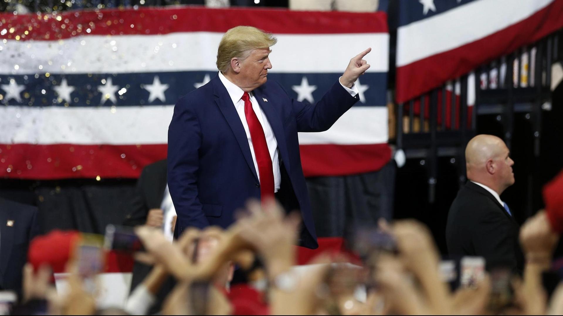 Trump steht auf einer Bühne vor einer Flagge der Republikaner und zeigt mit dem Zeigefinger auf jemanden. Im Vordergrund unscharf viele erhobene Hände seiner Anhänger.