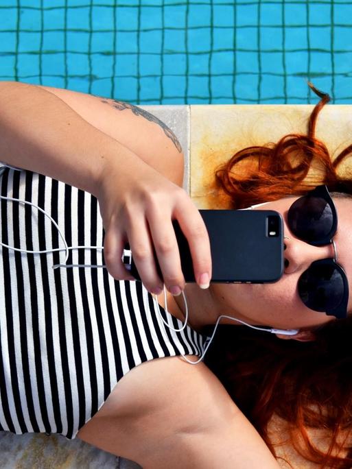 Eine junge Frau liegt am Rand eines Pools und kommunziert über ihr Handy.