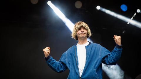 Der Musiker Tim Burgess steht in einer blauglänzenden Jacke auf einer Bühne, im Hintergrund Scheinwerferlicht.