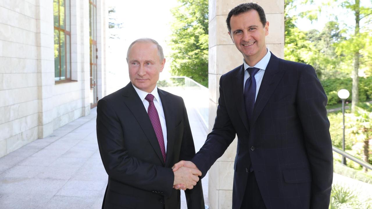 Wladimir Putin und Baschar al-Assad geben sich die Hand und lächeln in die Kamera