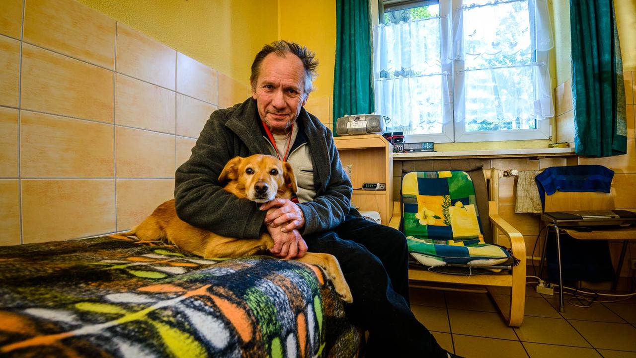 Ein ehemaliger Obdachlose sitzt zusammen mit einem Hund im Arm auf einem Bett in einem Zimmer des William-Booth-Haus der Heilsarmee in Berlin.
