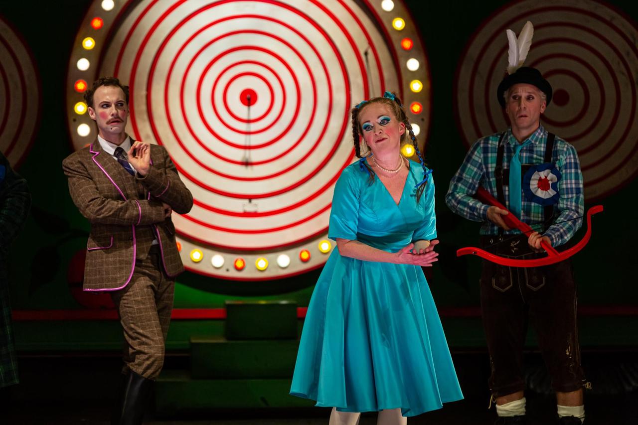 Eine Opernszene mit einer stilisierten Zielscheibe im Hintergrund. Vorne steht eine Frau im türkisfarbenen Kleid, rechts von ihr ein Mann mit Armbrust, links von ihr ein andere Mann im Anzug.