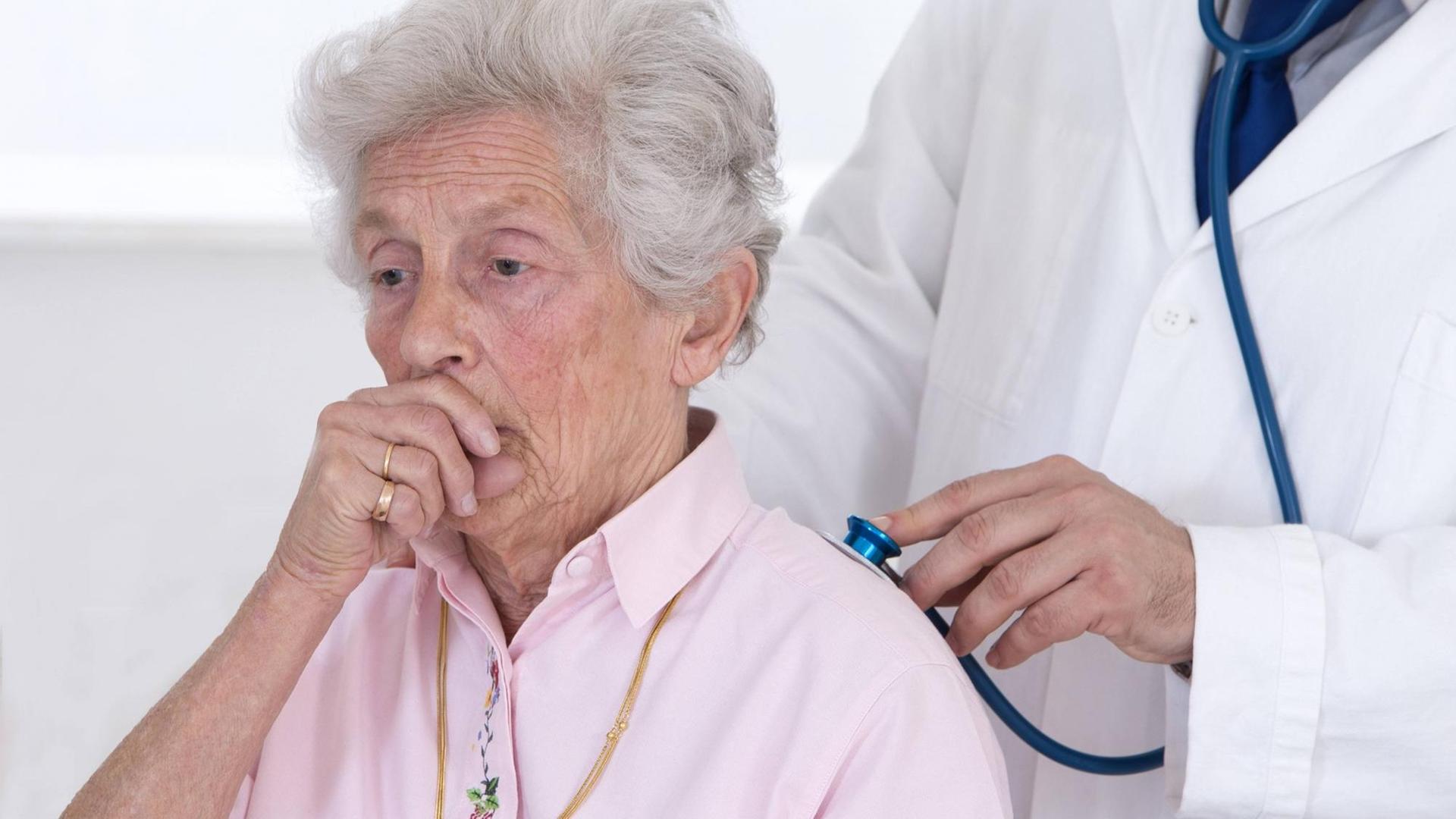 Eine ältere Frau wird mit einem Stethoskop am Rücken abgehört und hustet dabei in ihre Hand.