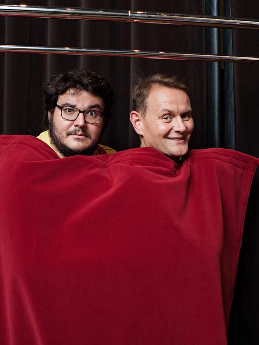 Das Bild zeigt Axel Ranisch und Devid Striesow, die sich zusammen in einen roten Vorhang eingewickelt haben.