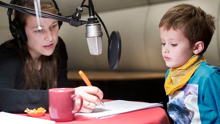 Eine Frau mit einem Kopfhörer auf dem Kopf und einem Kugelschreiber in der Hand sitzt gemeinsam mit einem kleinen Jungen vor einem Mikrofon im Tonstudio.