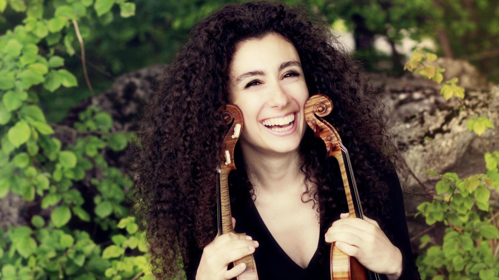 Eine lachende junge Frau mit langen schwarzen lockigen Haaren hält zwei Violinen in den Händen.