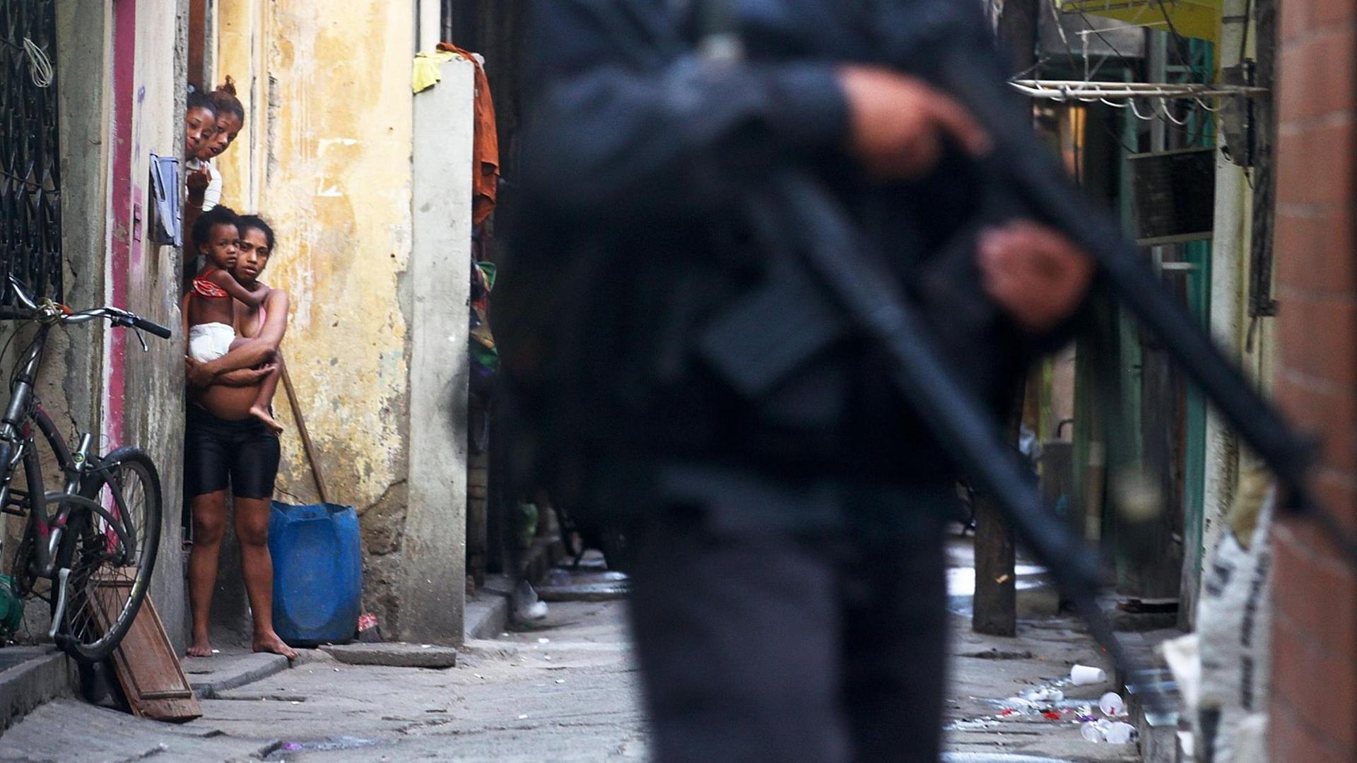 In der Favela Maré in der brasilianische Rio de Janeiro schauen drei schwarze Frauen und ein Kind aus einem Hauseinagang auf einen mit einem Gewehr bewaffenten Polizisten.