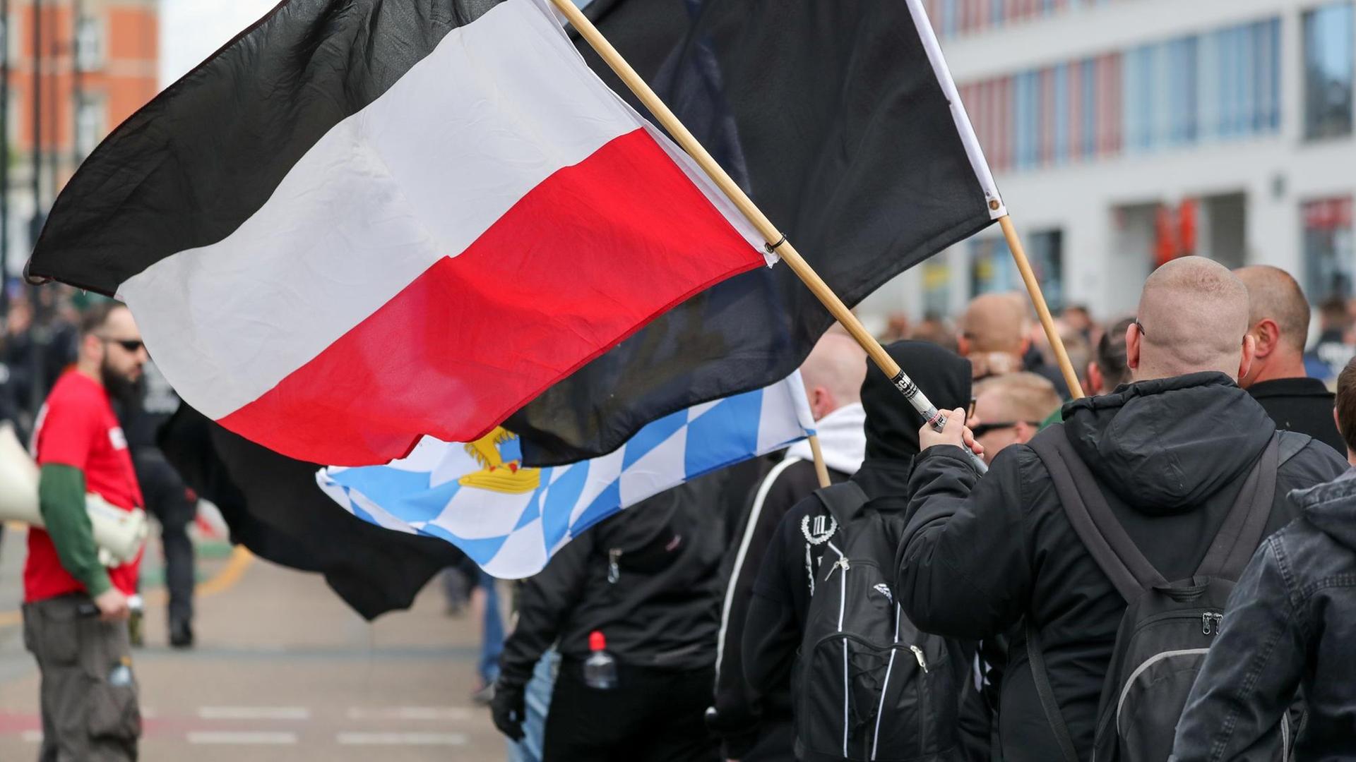 Glatzköpfige, in schwarz gekleidete Männer sind von hinten zu sehen, wie sie mit Flaggen durch die Straße ziehen.