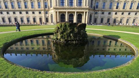 Spaziergänger gehen am 27.12.2015 im Hofgarten der Residenz in Würzburg (Bayern) hinter einem Brunnen entlang, in dem sich Teile der Residenz spiegeln