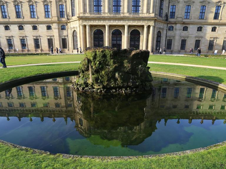 Spaziergänger gehen am 27.12.2015 im Hofgarten der Residenz in Würzburg (Bayern) hinter einem Brunnen entlang, in dem sich Teile der Residenz spiegeln.