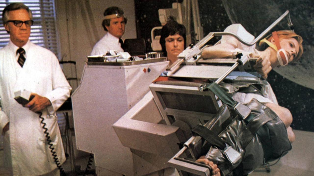 David Bowie in "Der Mann, der vom Himmel" festgebunden auf einem Laborstuhl in Schräglage. Im Hintergrund sind drei Wissenschaftler in weißen Kitteln zu sehen. 
