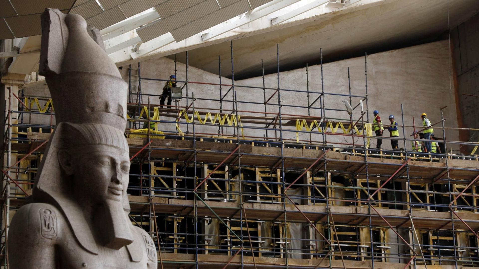 Auf der Baustelle des Großen Ägyptischen Museums in Kairo. Auf einem Baugerüst in großer Höhe stehen Bauarbeiter, links im Vordergrund steht eine riesige Statue eines Pharaos.
