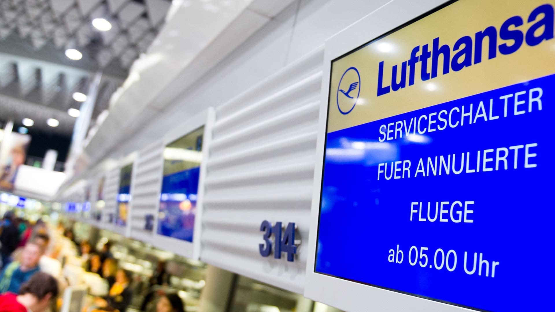 An einem Flugschalter der Lufthansa in Frankfurt am Main ist auf dem Display zu lesen: "Serviceschalter für annulierte Flüge ab 5 Uhr." Vor dem Schalter sind unscharf Fluggäste zu erkennen; dahinter sitzen zwei Service-Angestellte der Lufthansa.