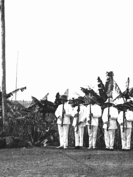 Auf einem Militärstützpunkt in der deutschen Kolonie Kamerun wird von in Tropenanzüge gekleideten Männern eine Fahne gehisst (undatierte Aufnahme aus der Kolonialzeit). Von 1884 bis zum Ersten Weltkrieg war Kamerun eine deutsche Kolonie, dann wurde es 1916 unter Großbritannien und Frankreich aufgeteilt.