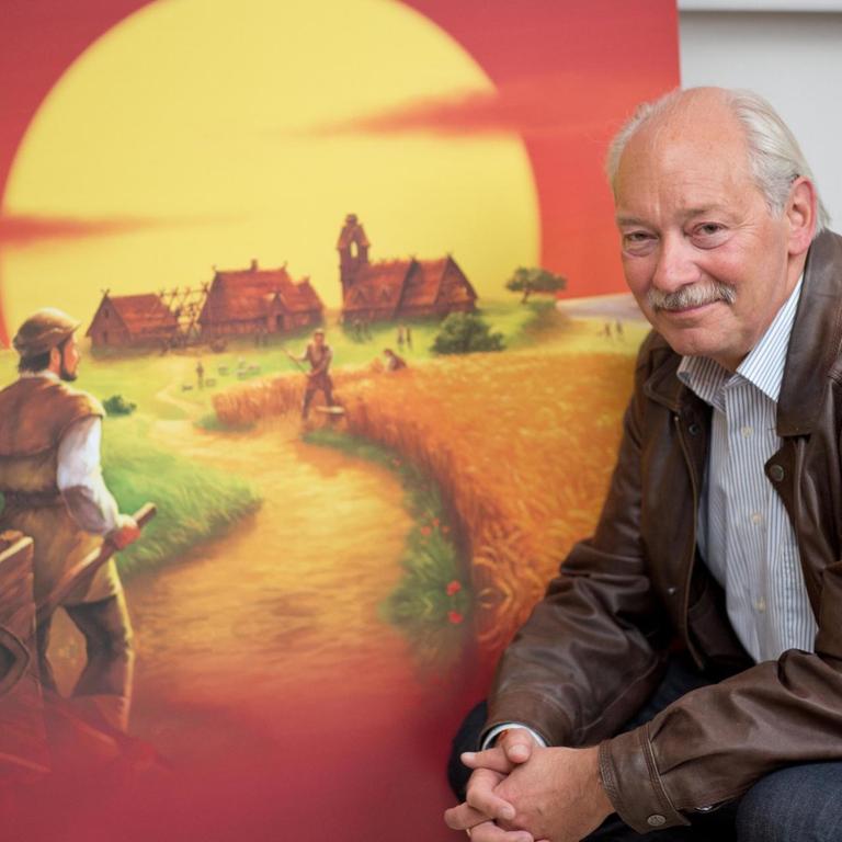 Klaus Teuber sitzt vor dem Bild seines Spiels "Die Siedler von Catan"