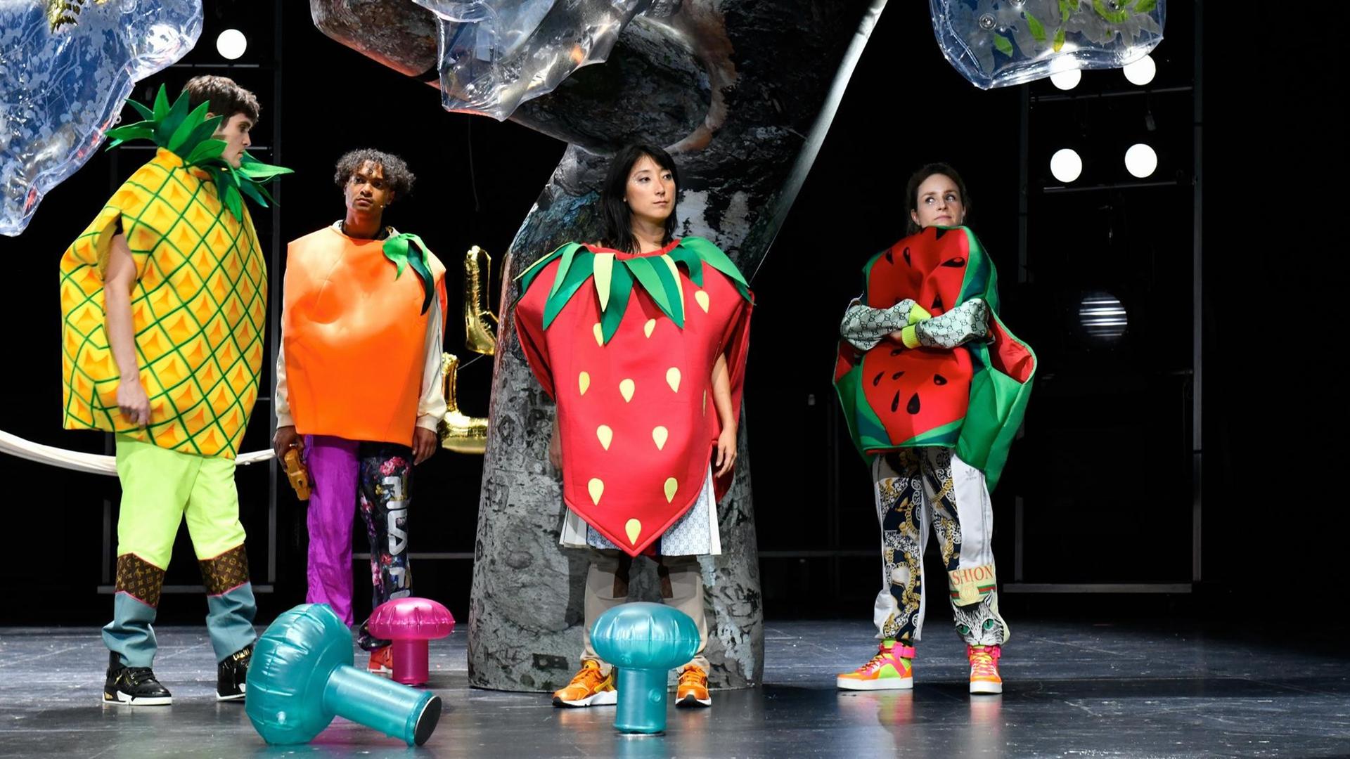Vier Schauspieler stehen als Früchte wie Erdbeere und Ananas verkleidet auf einer Theaterbühne.