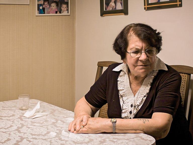 Die Shoah-Überlebende Frieda Kliger sitzt an ihrem Küchentisch und blickt nachdenklich. Auf Ihrem linken Arm ist eine tätowierte Nummer erkennbar.