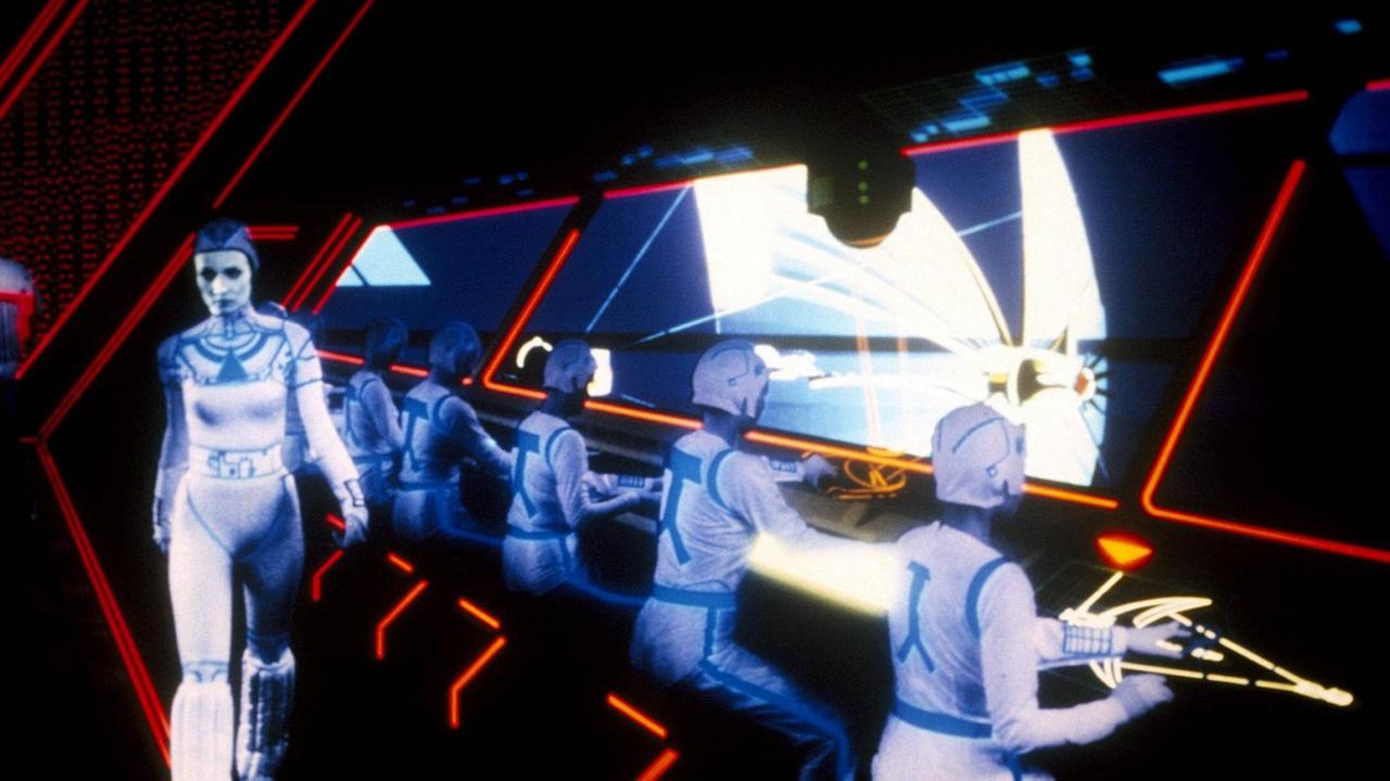 Im Still aus "Tron" sitzen uniformierte Darsteller an einer futuristisch anmutenden Konsole.
