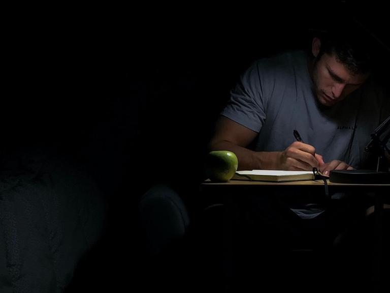 Ein Mann sitzt in einem dunklen Zimmer und schreibt im Schein einer zielgerichteten Lampe mit der Hand in einen Block,