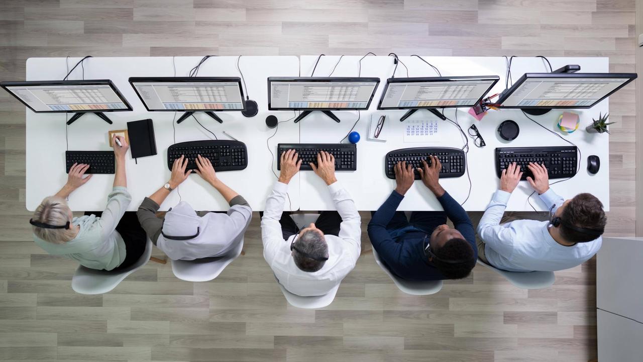 Draufsicht auf einen weißen Schreibtisch mit diversen Computerbildschirmen vor denen eng nebeneinander fünf Menschen arbeiten