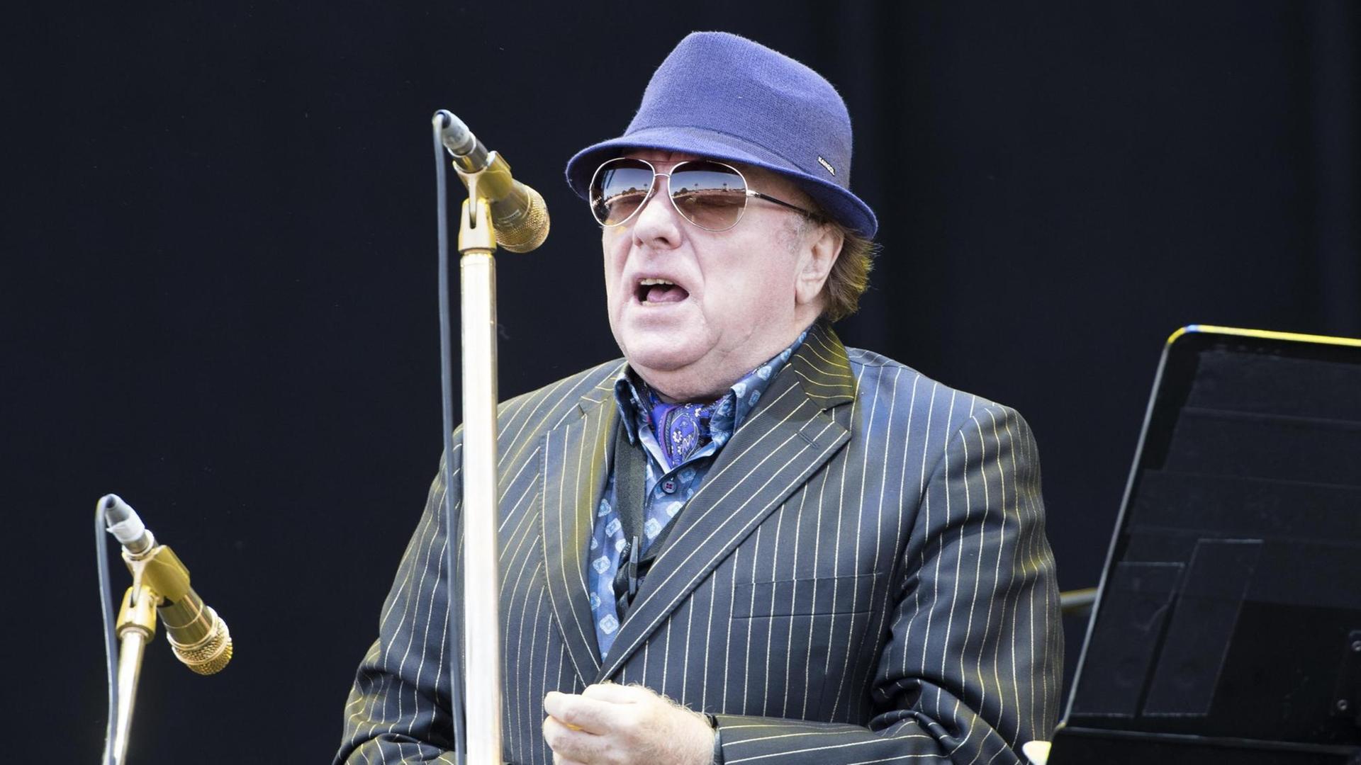 Der irische Musiker Van Morrison steht in Anzug und Hut auf einer Bühne, vor ihm ein goldfarbenes Mikrofon.