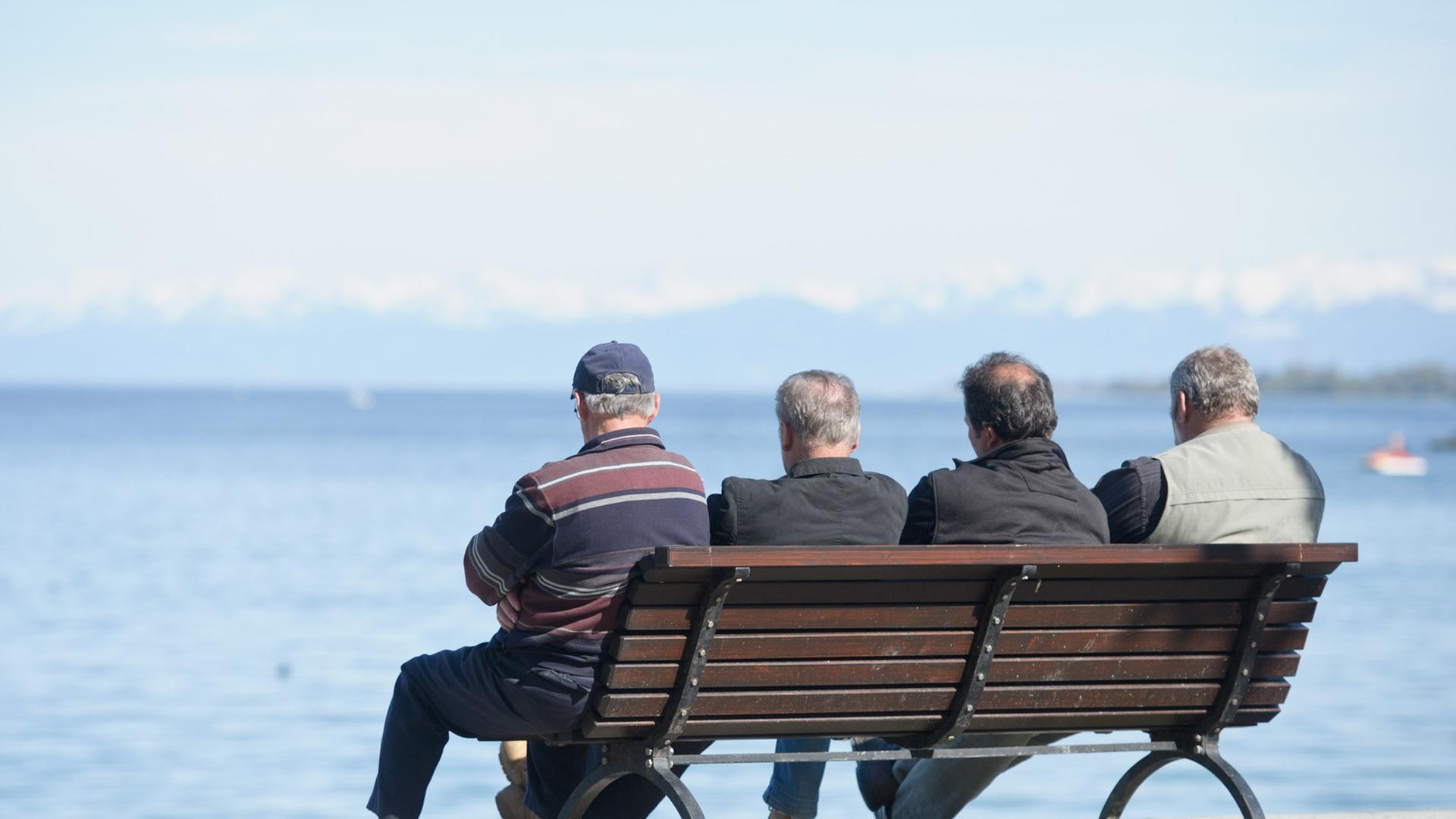 Vier Herren fortgeschrittenen Alters sitzen auf einer Bank am Ufer und blicken auf das Wasser.