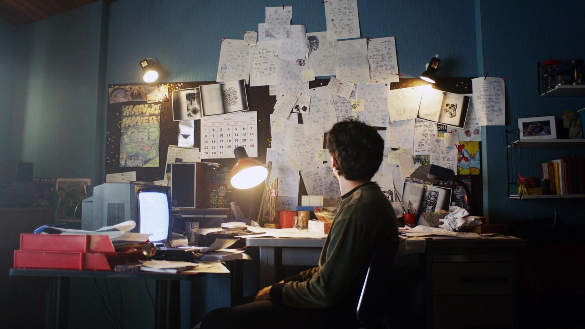 Filmstill aus der Netflix Serie "BLACK MIRROR: BANDERSNATCH. Der Schauspieler Fionn Whitehead in der Hauptrolle sitzt vor einem Bildschirm an einem chaotischem Schreibtisch.