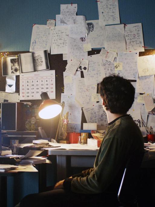 Filmstill aus der Netflix Serie "BLACK MIRROR: BANDERSNATCH. Der Schauspieler Fionn Whitehead in der Hauptrolle sitzt vor einem Bildschirm an einem chaotischem Schreibtisch.